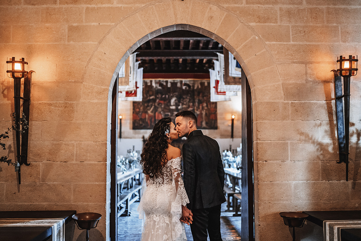 Pareja de novios besándose bajo un arco de piedra del Castillo de Almodóvar, con el salón iluminado con antorchas al fondo